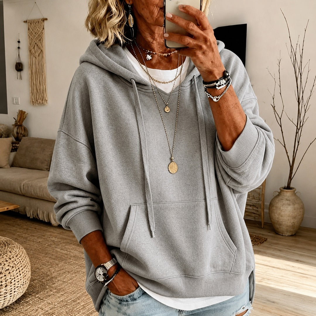 Vrouw in een lichtgrijze oversized trui met capuchon over een wit topje en een lichte jeans, in de woonkamer | Boutique Moda
