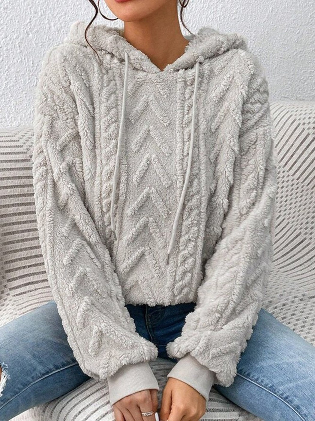 Vrouw in een lichtgrijze teddytrui met zigzag- en kabelpatroon, zittend op een bank | Boutique Moda