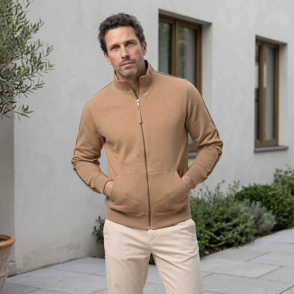 Man in een camelkleurig sweatvest op een binnenplaats voor een lichte gevel, met beide handen in de zakken | Boutique Moda