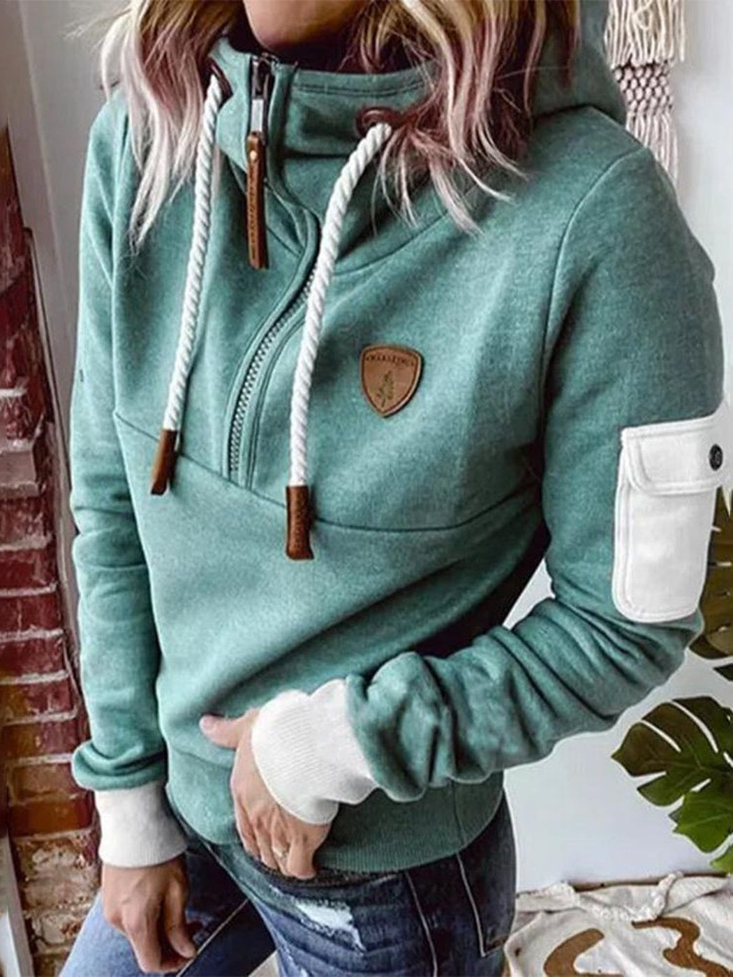 Vrouw in een mintgroene hoodie met hoge kraag, wit mouwzakje en bruine patch, hand in de zak van haar jeans | Boutique Moda