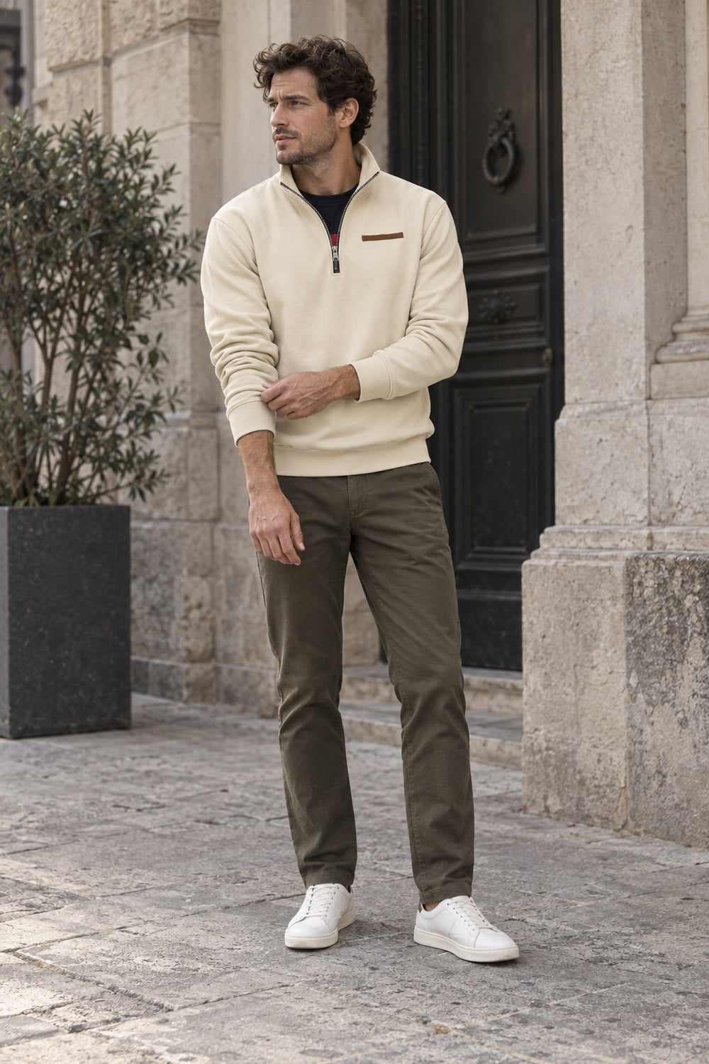 Man ten voeten uit in een beige sweater met een olijfgroene broek en witte sneakers voor een stenen gevel | Boutique Moda