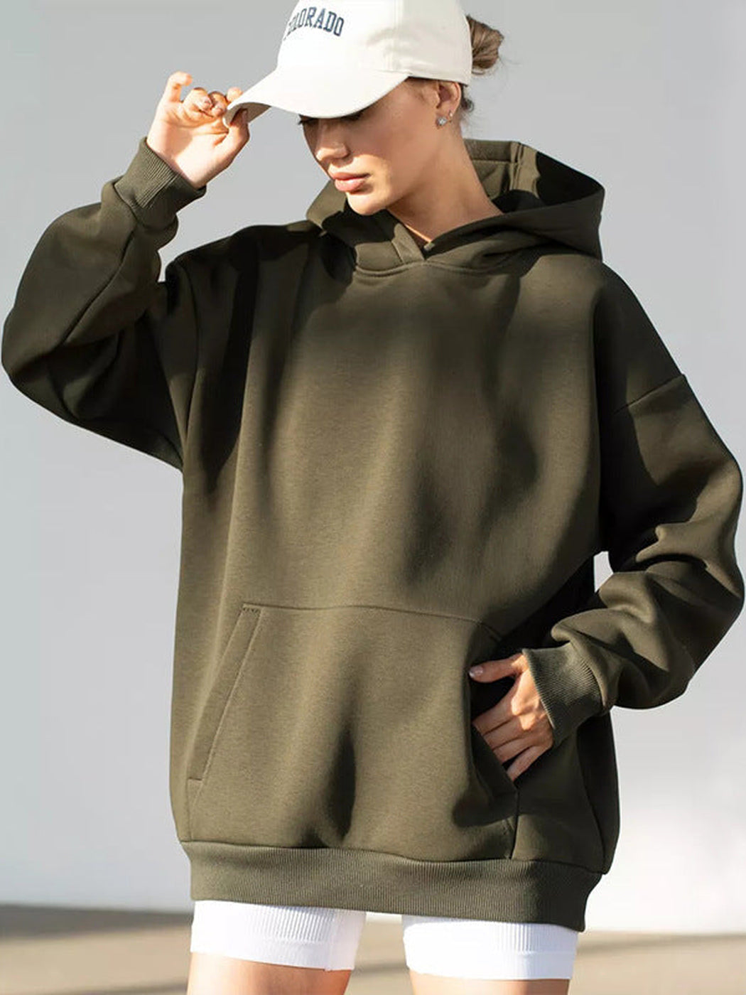 Vrouw met een lichte pet in een olijfgroene hoodie, hand aan de pet | Boutique Moda