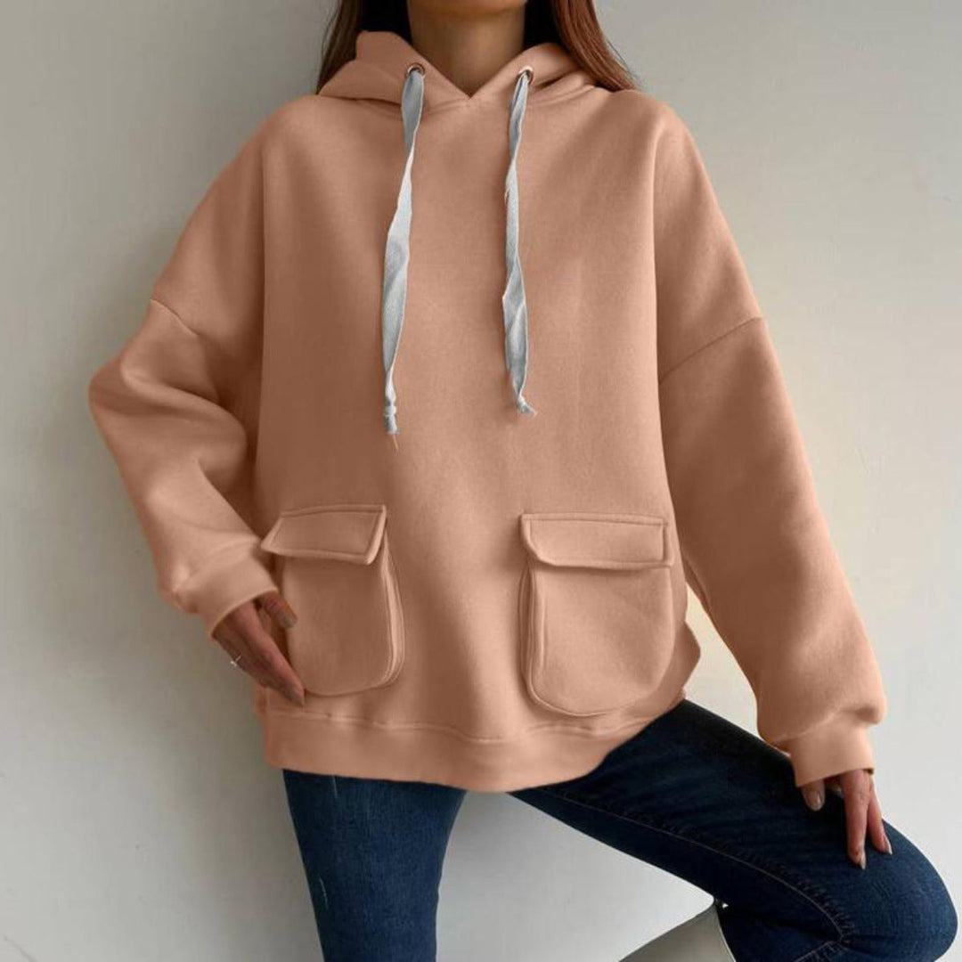 Vrouw in een perzikkleurige oversized hoodie met lichtgrijze koorden en klepzakken, voor een grijze muur | Boutique Moda