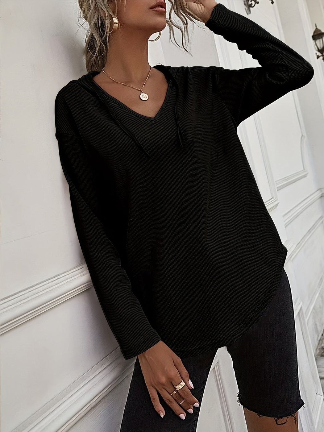 Vrouw in een zwarte hoodie met een zwarte short, leunend tegen een witte muur | Boutique Moda