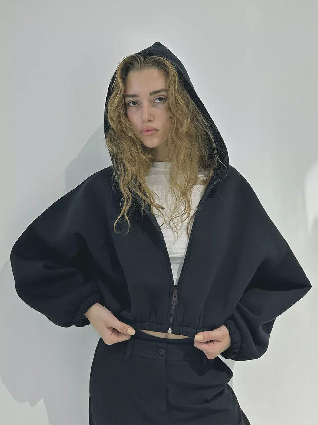 Vrouw in een zwart kort vest met opgezette capuchon, handen aan de elastische zoom | Boutique Moda
