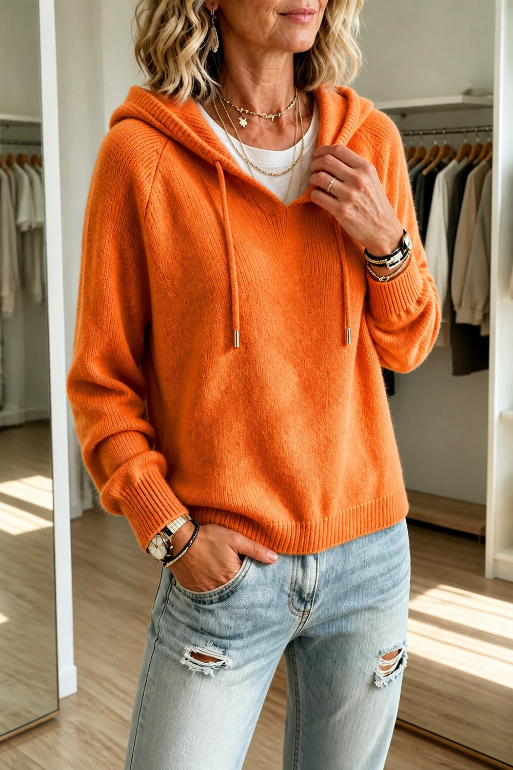 Vrouw in een oranje gebreide hoodie met raglanmouwen, hand bij de halsopening | Boutique Moda