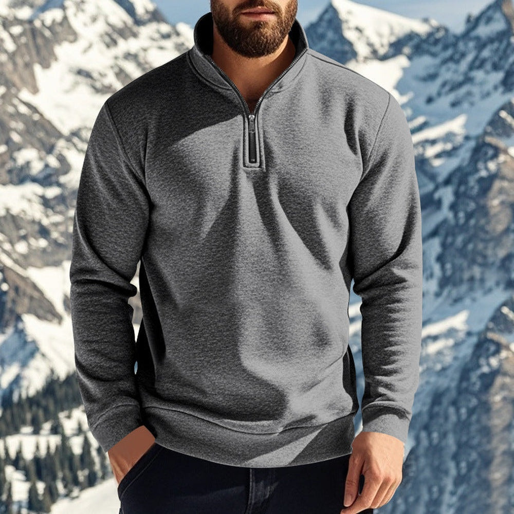 Man in een donkergrijze gevoerde trui met een hand in zijn broekzak, besneeuwde bergen op de achtergrond | Boutique Moda