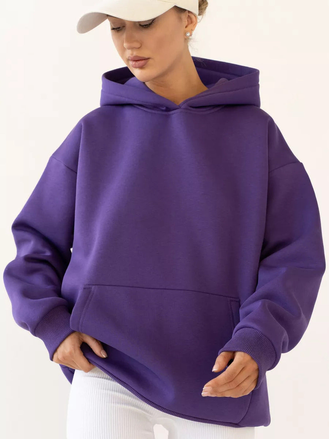 Vrouw met een lichte pet in een donkerpaarse hoodie, beide handen aan de zoom | Boutique Moda