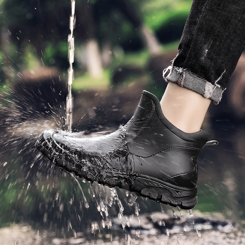 A black ankle rain boot being splashed with water, worn on a person's foot with rolled-up jeans.