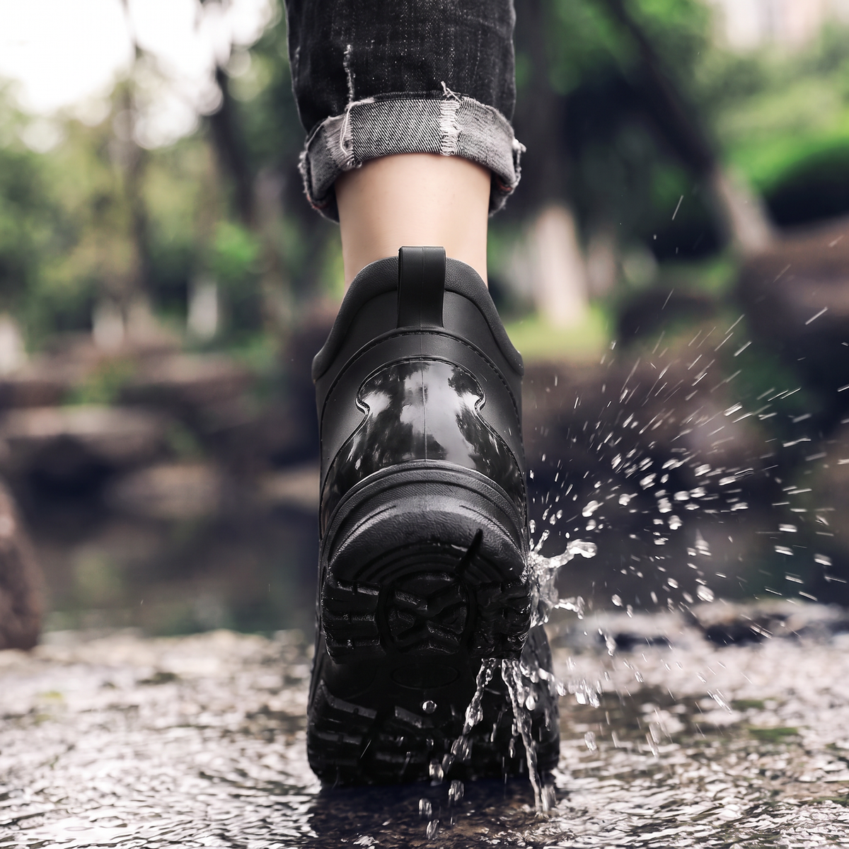 A black ankle rain boot being splashed with water, worn on a person's foot with rolled-up jeans, back view