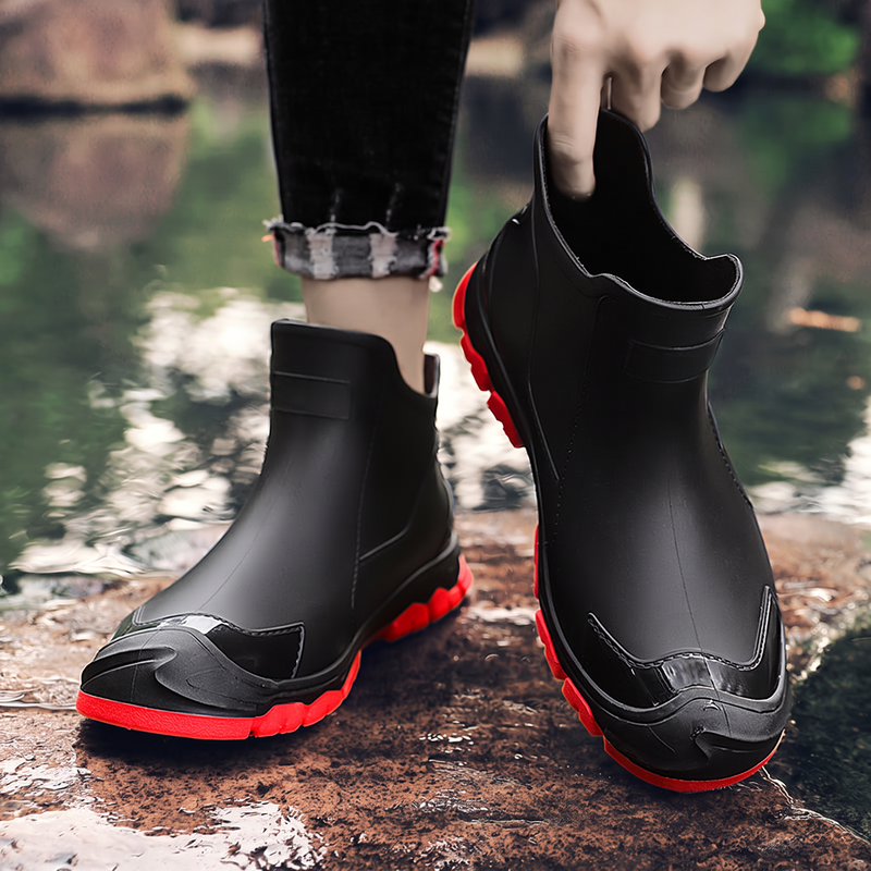 A pair of black ankle rain boots with red soles, one worn on a foot and one held by a hand, near water.