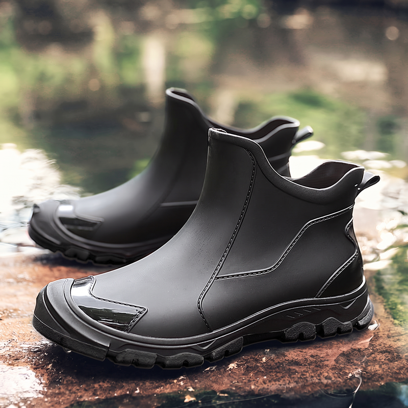 A pair of black ankle rain boots with textured soles displayed on a wet rocky surface near water.