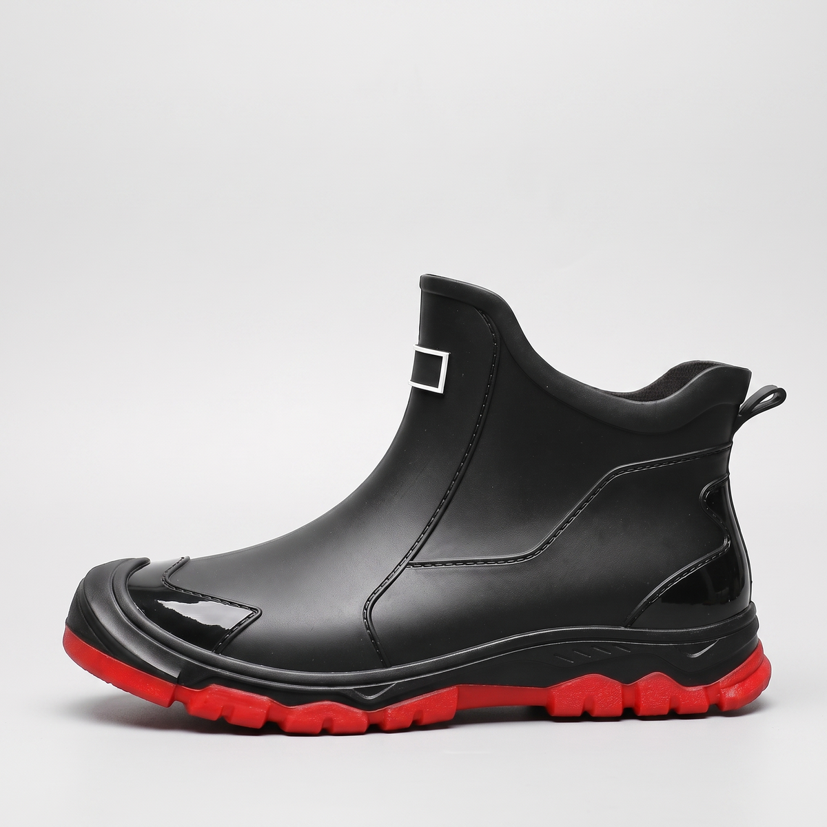 A black ankle rain boot with a red tread sole and a small buckle detail at the top, shown against a plain grey background.