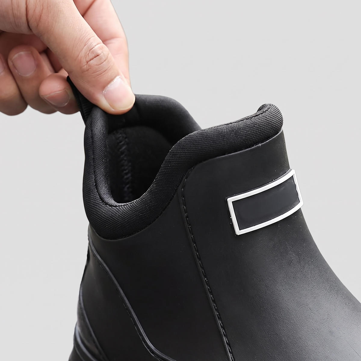 A hand pulls back the top collar of a black ankle rain boot, showing a rectangular metal buckle detail near the heel.