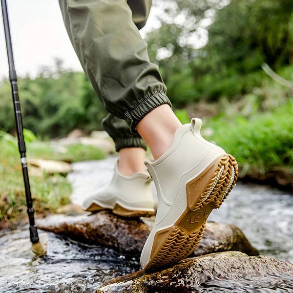 A person in olive cargo trousers wears cream ankle rain boots with tan treaded soles while stepping across rocks in a stream, with a trekking pole beside them.