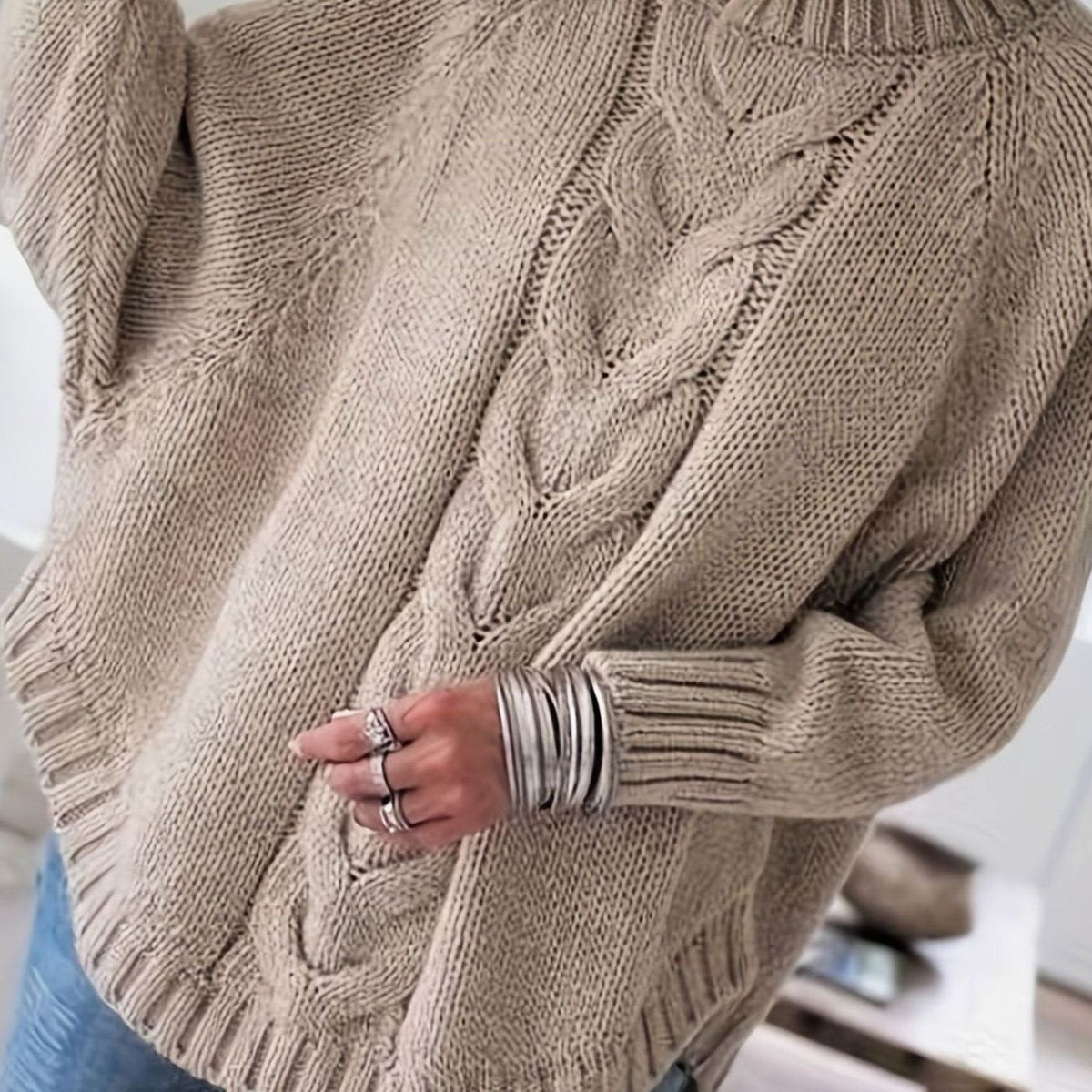 A close up of a person's back and arm wearing an oversized beige cable knit sweater, with the hand showing rings and bracelets.