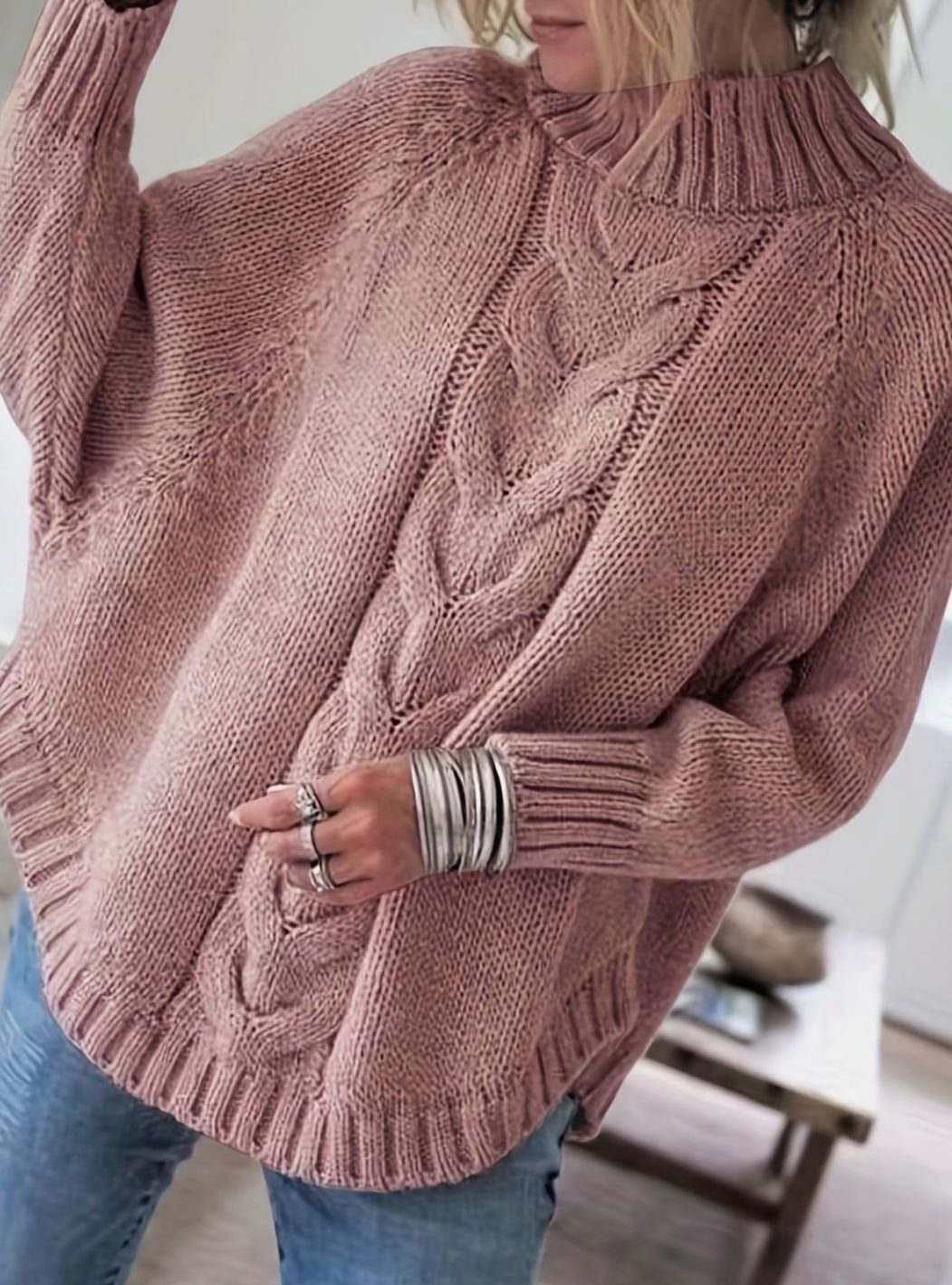 A woman wearing an oversized pink cable knit mock neck sweater with jeans, holding a hand up to her hair.