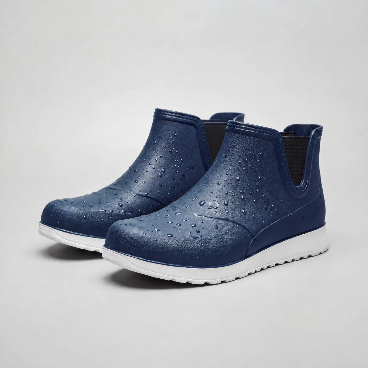 A pair of navy blue rubber ankle boots with water droplets on the surface and white chunky soles, photographed on a plain light background.