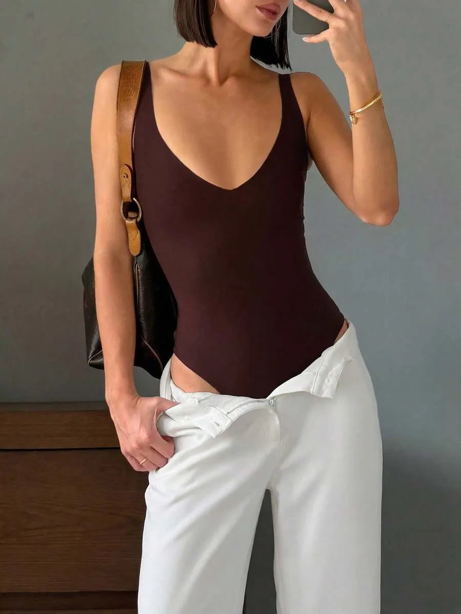A woman wears a deep V-neck brown bodysuit with white trousers while carrying a brown shoulder bag and holding a phone.