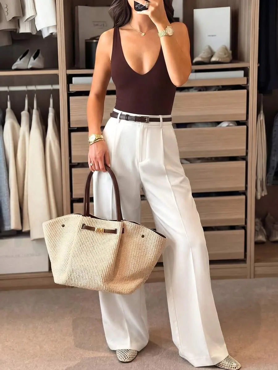 A woman in a closet wears a brown deep V-neck bodysuit with white wide-leg trousers and a belt, holding a woven tote bag.