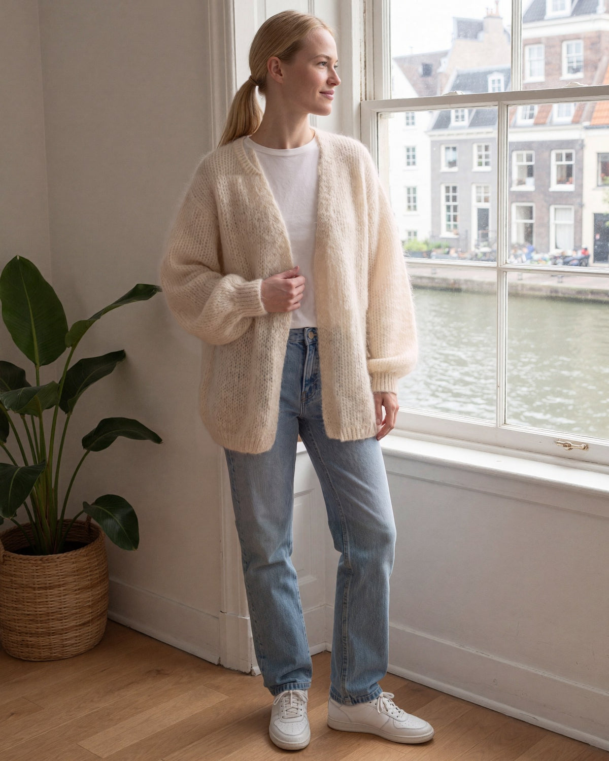 Vrouw draagt een beige oversized gebreid vest met ballonmouwen over een wit T-shirt en jeans, bij het raam van een grachtenpand | Boutique Moda