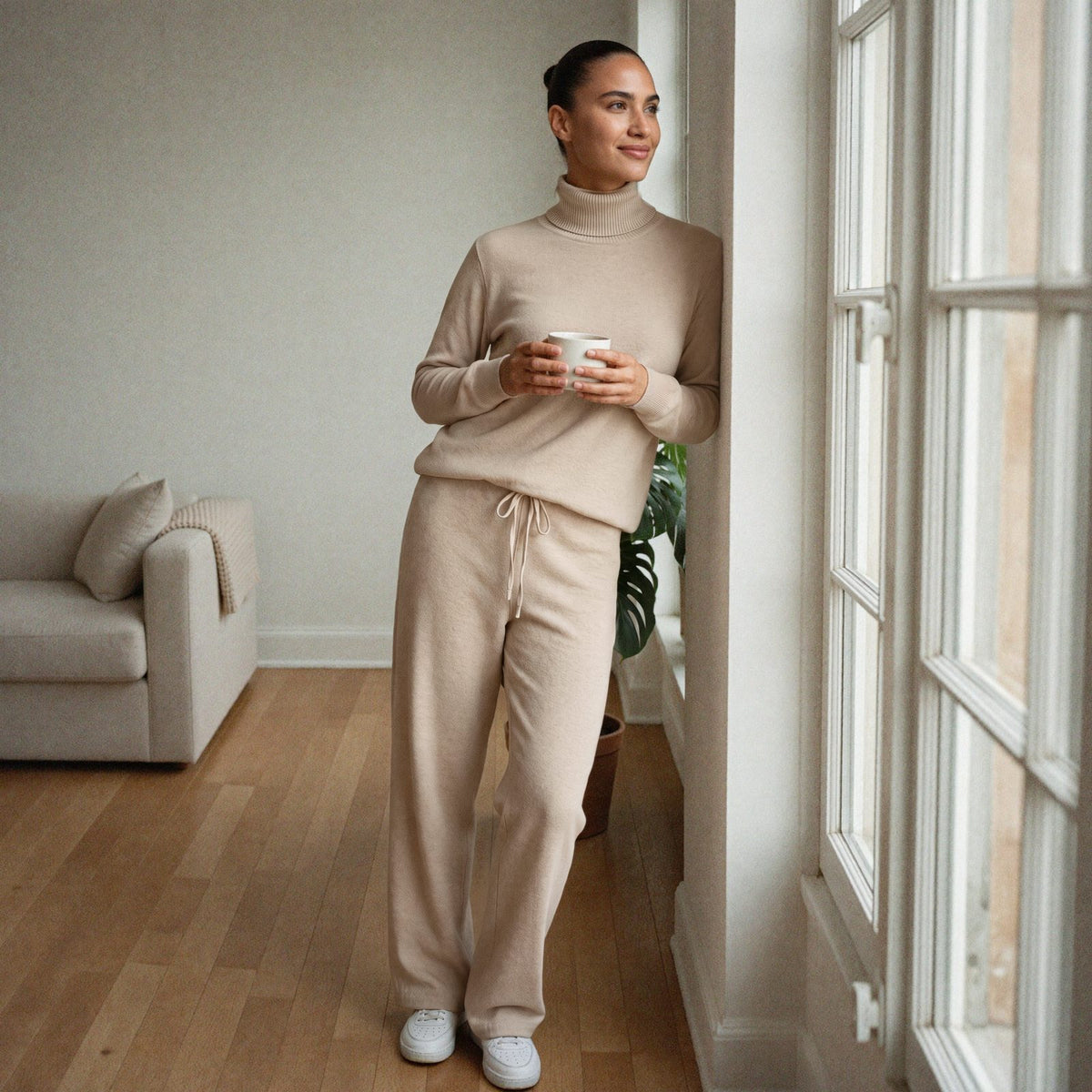Vrouw draagt een beige gebreid huispak met col en wijde broek bij het raam in een lichte woonkamer | Boutique Moda