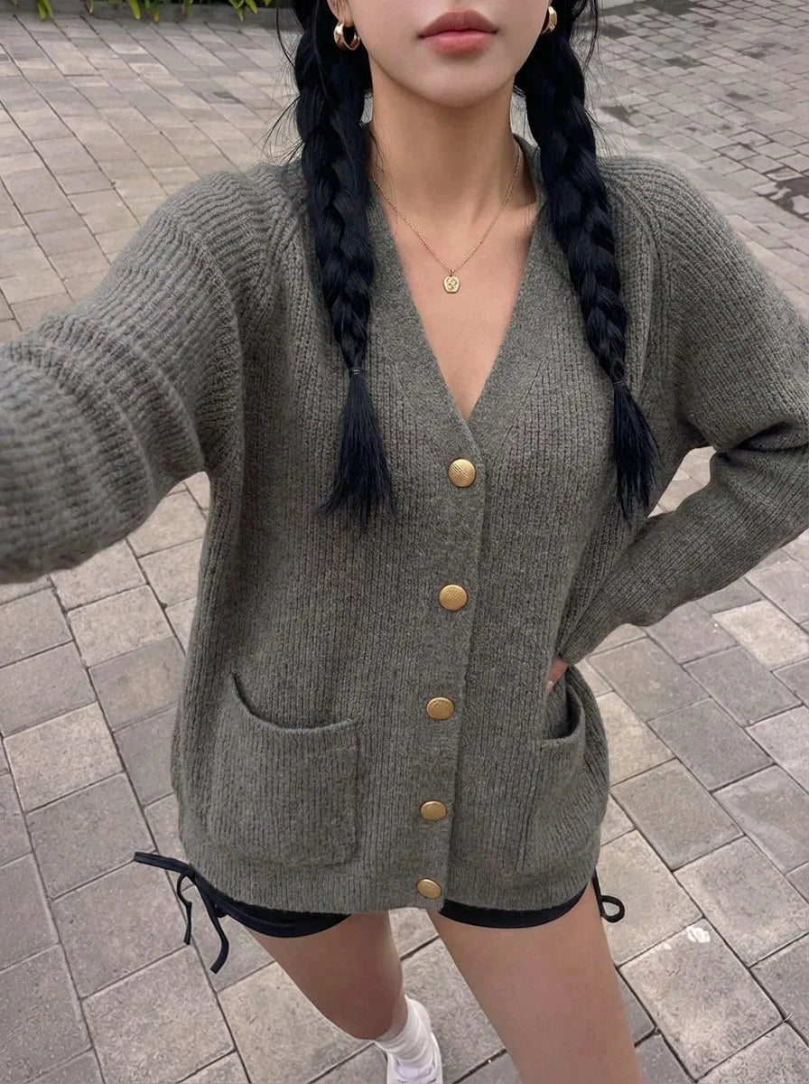 A woman wearing a gray oversized knit cardigan with gold buttons and a pendant necklace, standing on a cobblestone street.
