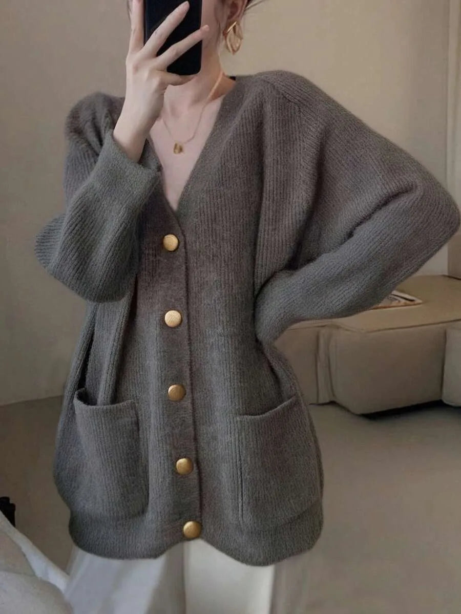 A woman wearing an oversized grey knit cardigan with gold buttons and patch pockets, taking a mirror selfie with a phone.