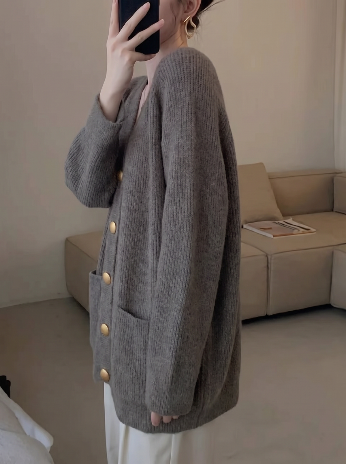 A woman wearing an oversized grey knit cardigan with gold buttons and patch pockets, taking a mirror selfie with a phone, side view