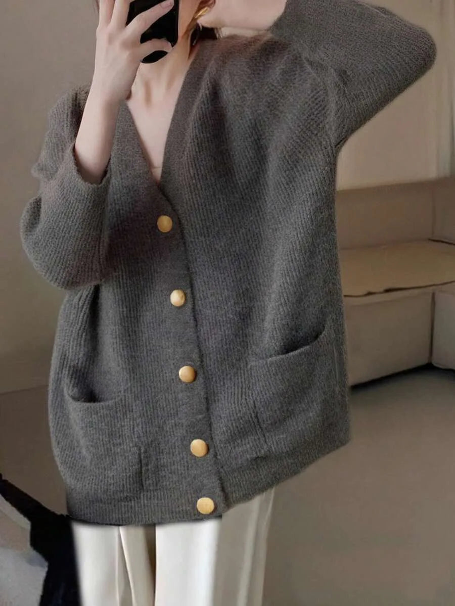 A woman wears an oversized grey knit cardigan with gold buttons and front pockets, holding a phone up to her face.