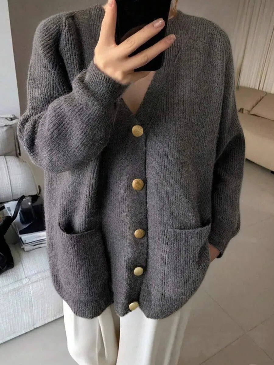 A woman wearing an oversized gray ribbed cardigan with gold buttons, holding a phone up to her face while taking a mirror selfie.