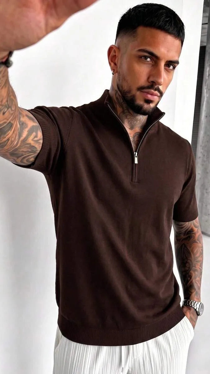 A man with tattoos and a watch takes a selfie wearing a brown knit half-zip polo shirt with white trousers.