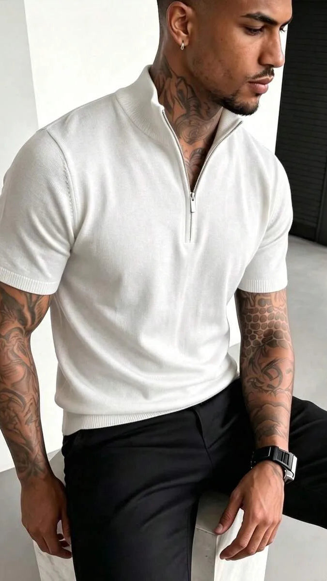 A man with tattooed arms and neck wears a white short sleeve half-zip knit polo with black trousers and a watch.