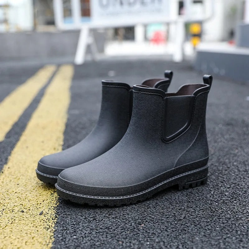A pair of black rubber Chelsea style rain boots with elastic side panels and pull tabs, standing on dark asphalt next to yellow road markings in a blurred street scene.