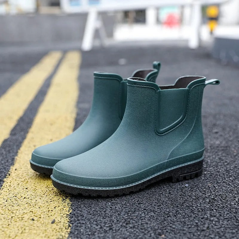 A pair of dark green Chelsea style rubber rain boots with black soles, standing on an asphalt street beside yellow road markings.