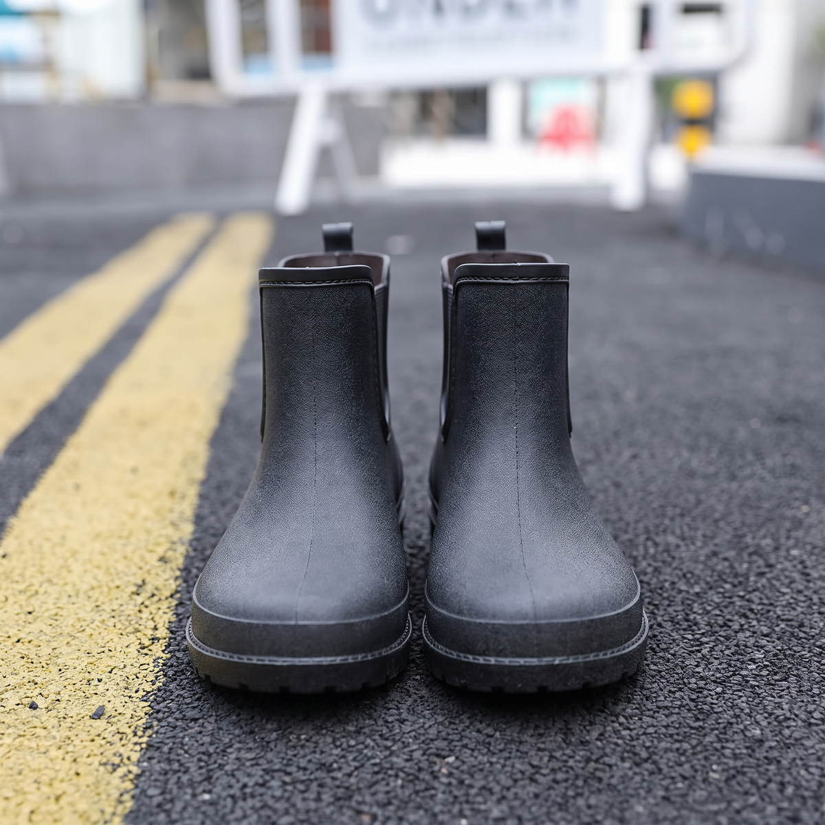 A pair of black ankle-height Chelsea style rubber rain boots with elastic side panels and pull tabs, standing on asphalt next to yellow road lines with a blurred street scene behind, front view