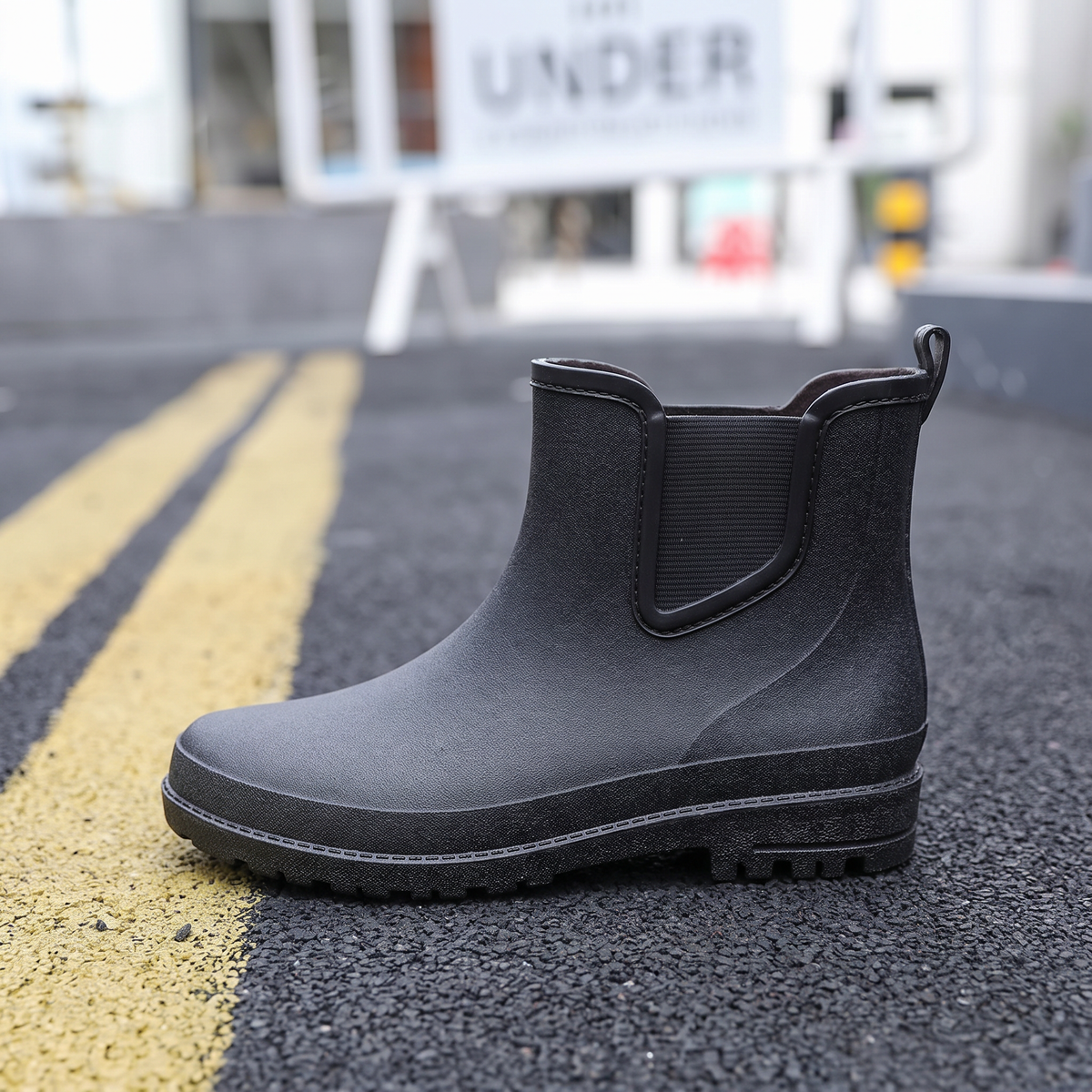 A pair of black ankle-height Chelsea style rubber rain boots with elastic side panels and pull tabs, standing on asphalt next to yellow road lines with a blurred street scene behind, side view
