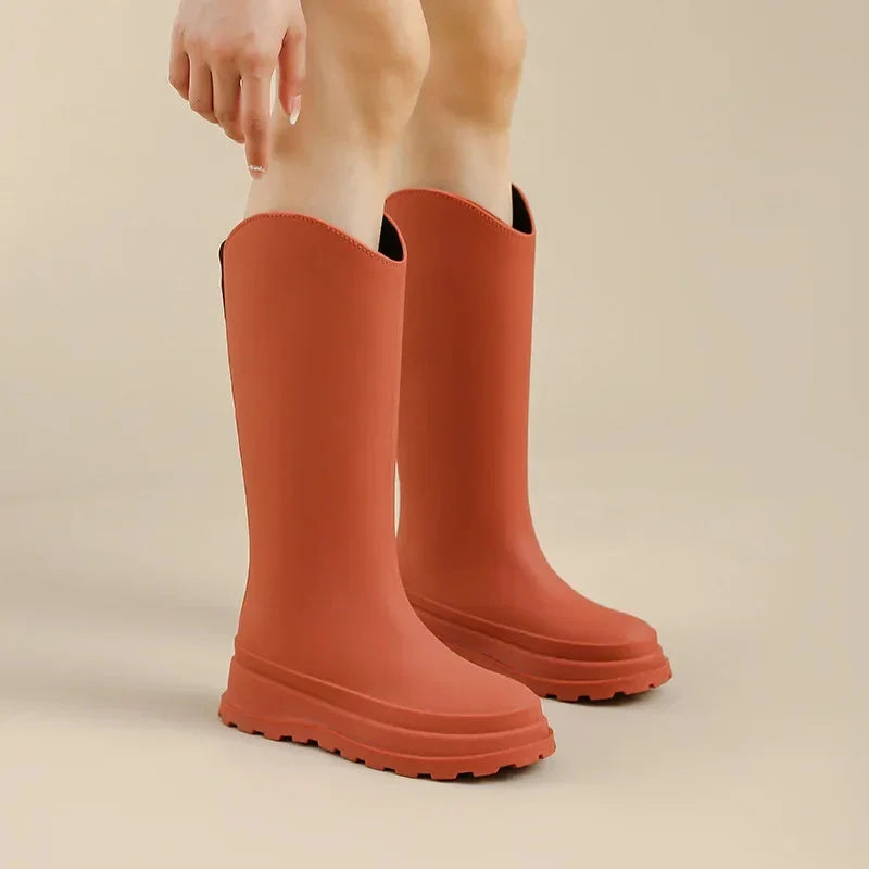 A woman's lower legs and feet wearing tall orange platform rain boots against a plain beige background.