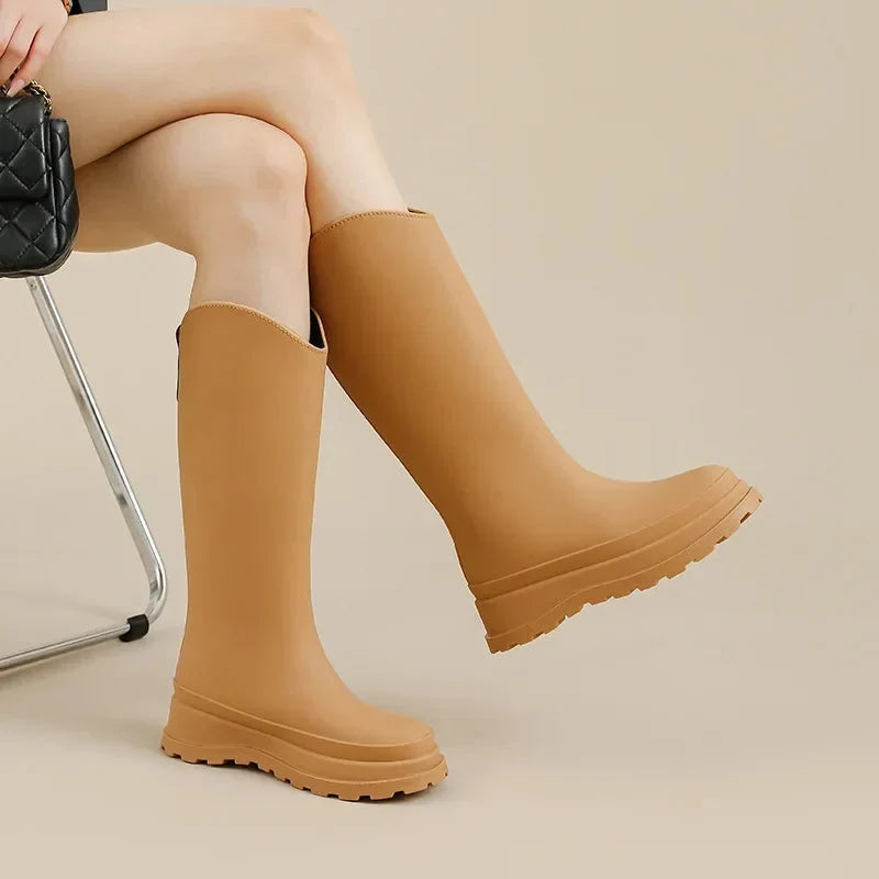A seated woman wears tan knee high platform rain boots while holding a quilted black handbag.