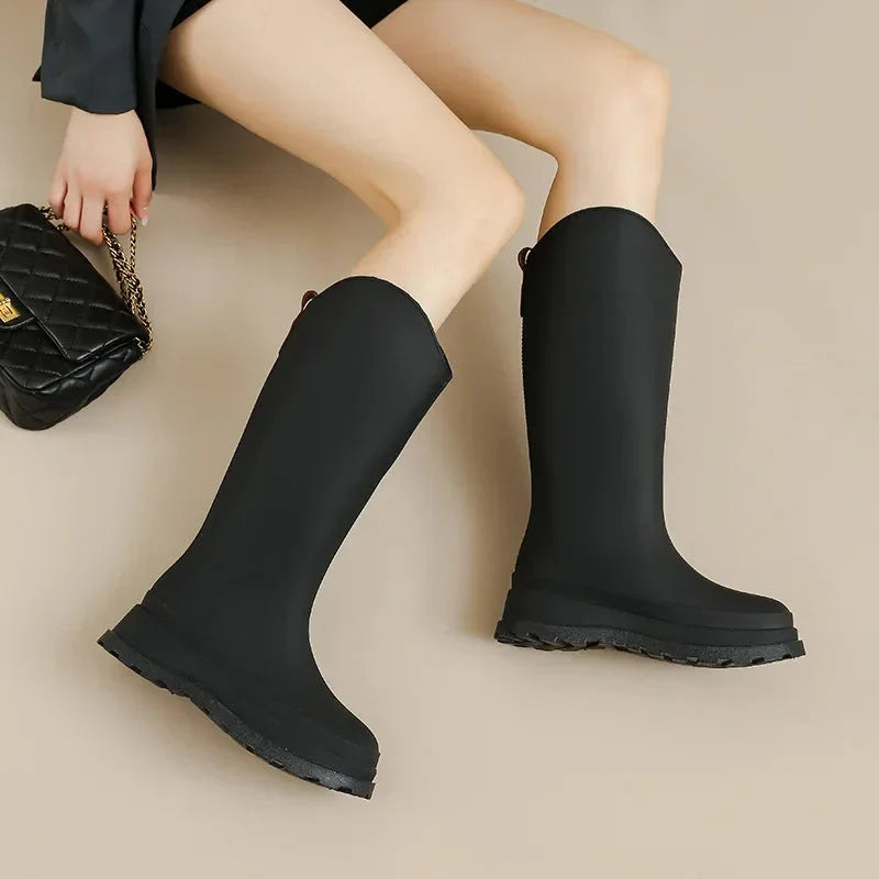 A woman sits on the floor wearing black knee high platform rain boots, holding a quilted black handbag.