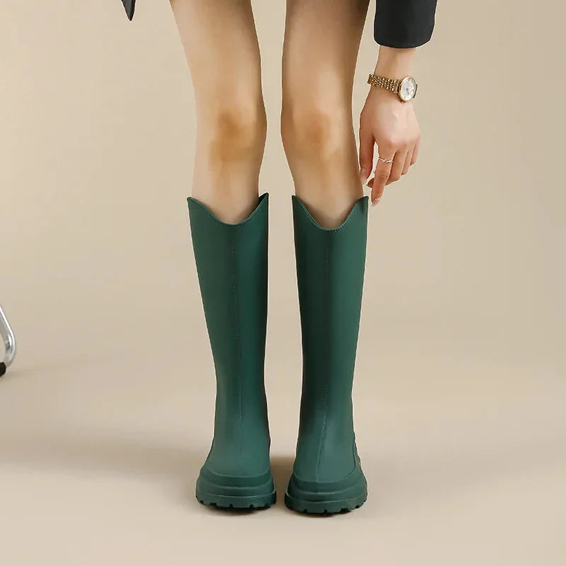 A woman wearing dark green tall rain boots with a chunky platform sole, shown from the knees down.