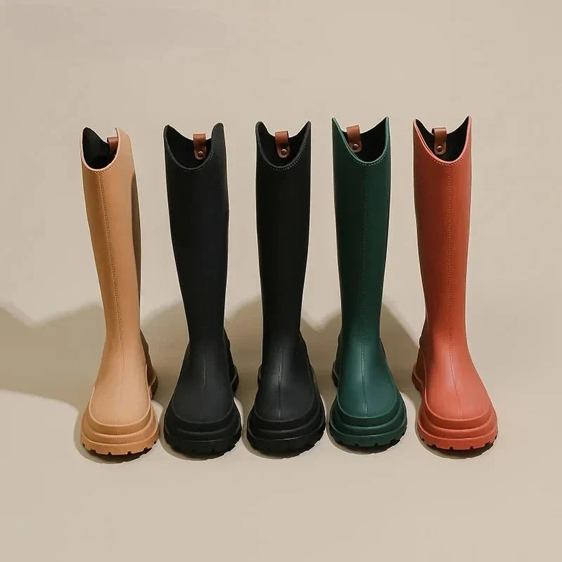 Five tall knee high rubber rain boots in tan, black, dark green and orange standing in a row against a plain beige background.