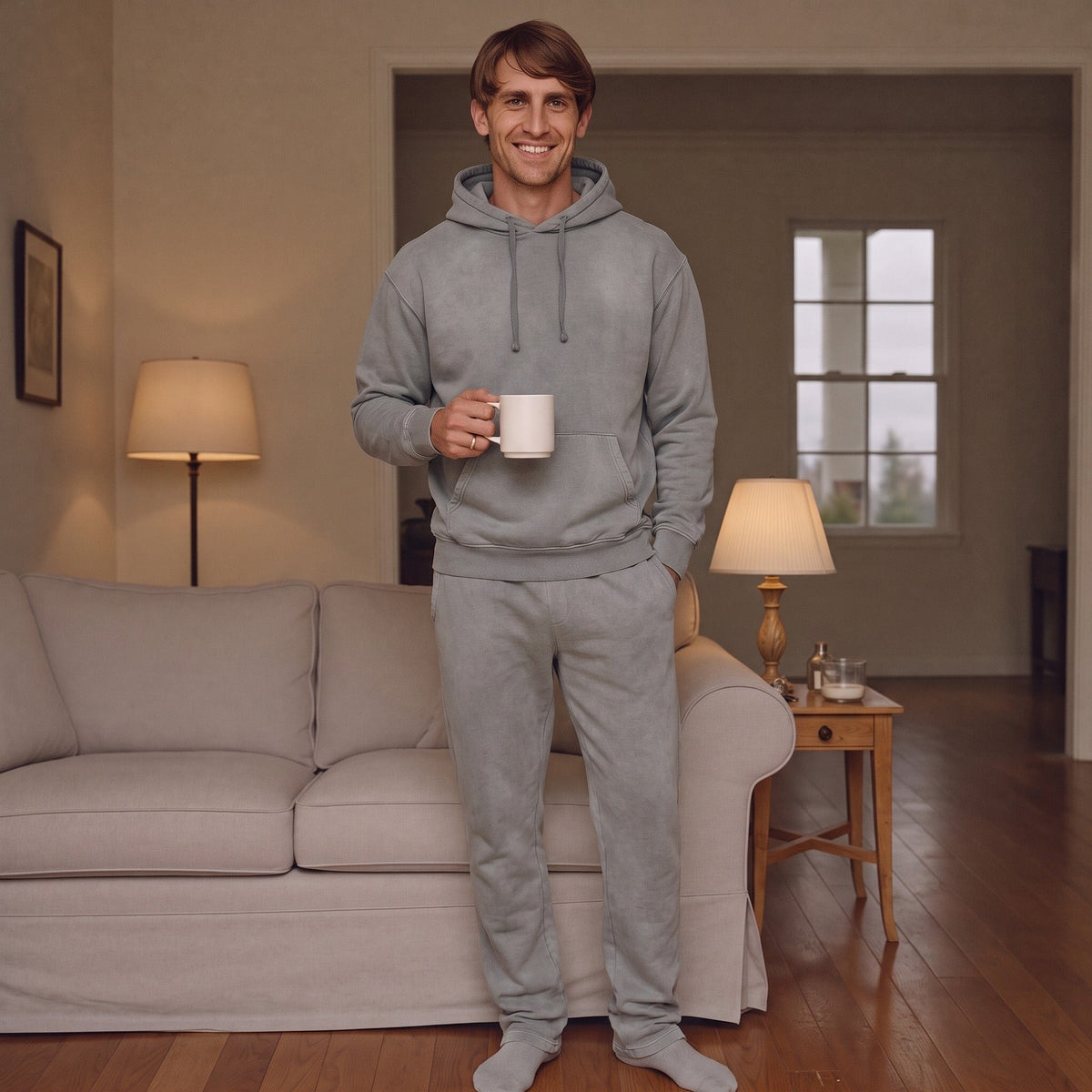 Man draagt grijze hoodie en joggingbroek, staand in woonkamer met koffiemok | Boutique Moda