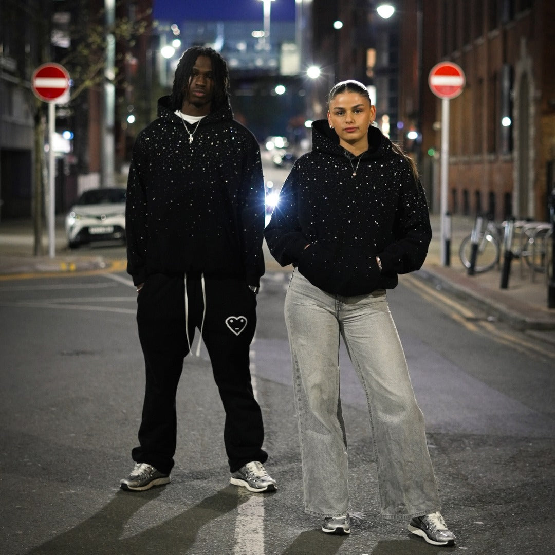 Zwarte oversized hoodie met strass, gedragen door een man en een vrouw, 's avonds in de stad