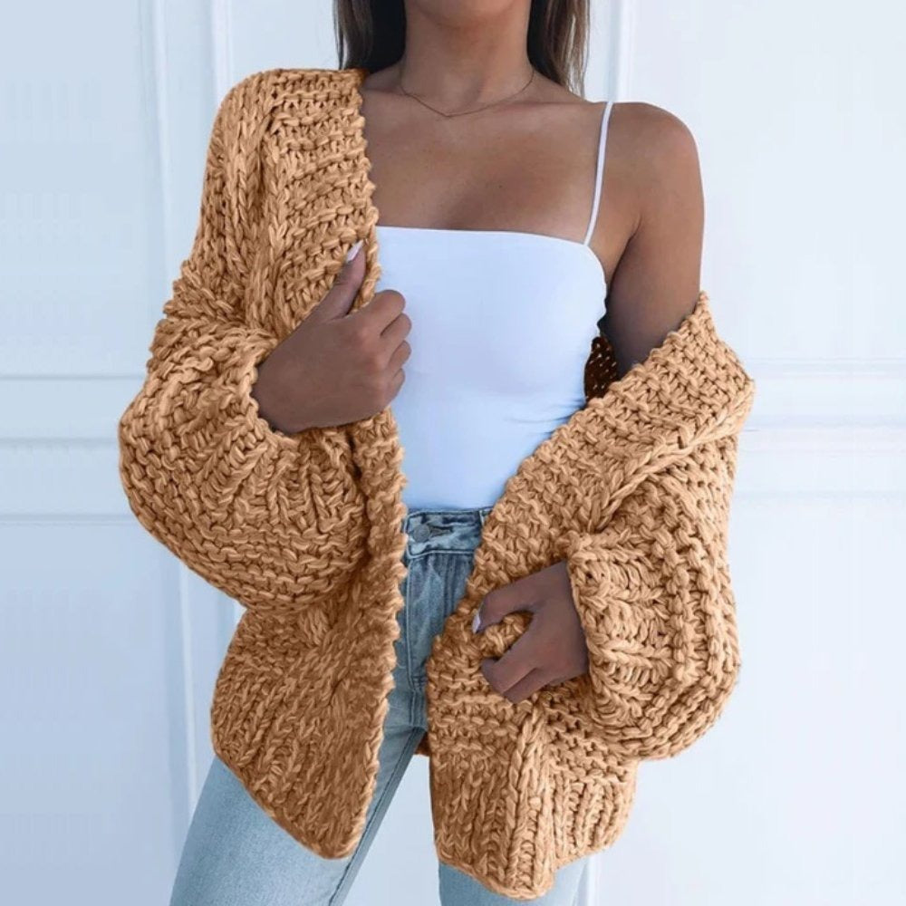 Grofgebreid oversized damesvest in camel, open gedragen over een wit topje, voor een witte muur met lijstwerk