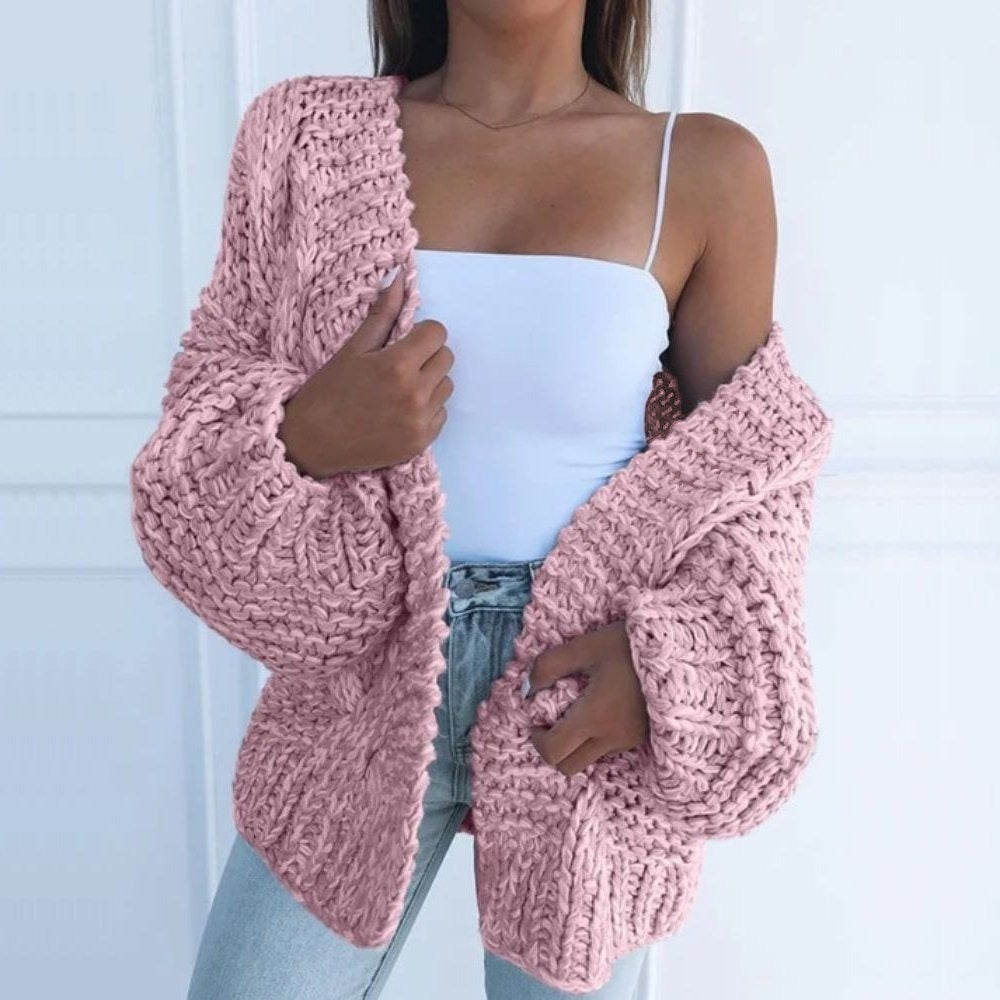 Grofgebreid oversized damesvest in roze, open gedragen over een wit topje, voor een witte muur met lijstwerk