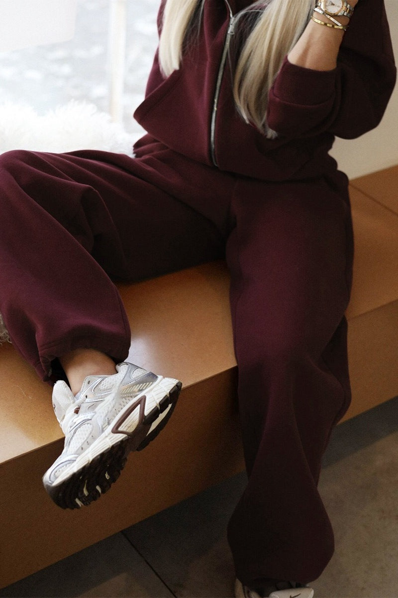Bordeaux joggingpak met capuchon, gedragen door zittende vrouw met sneakers | Boutique Moda