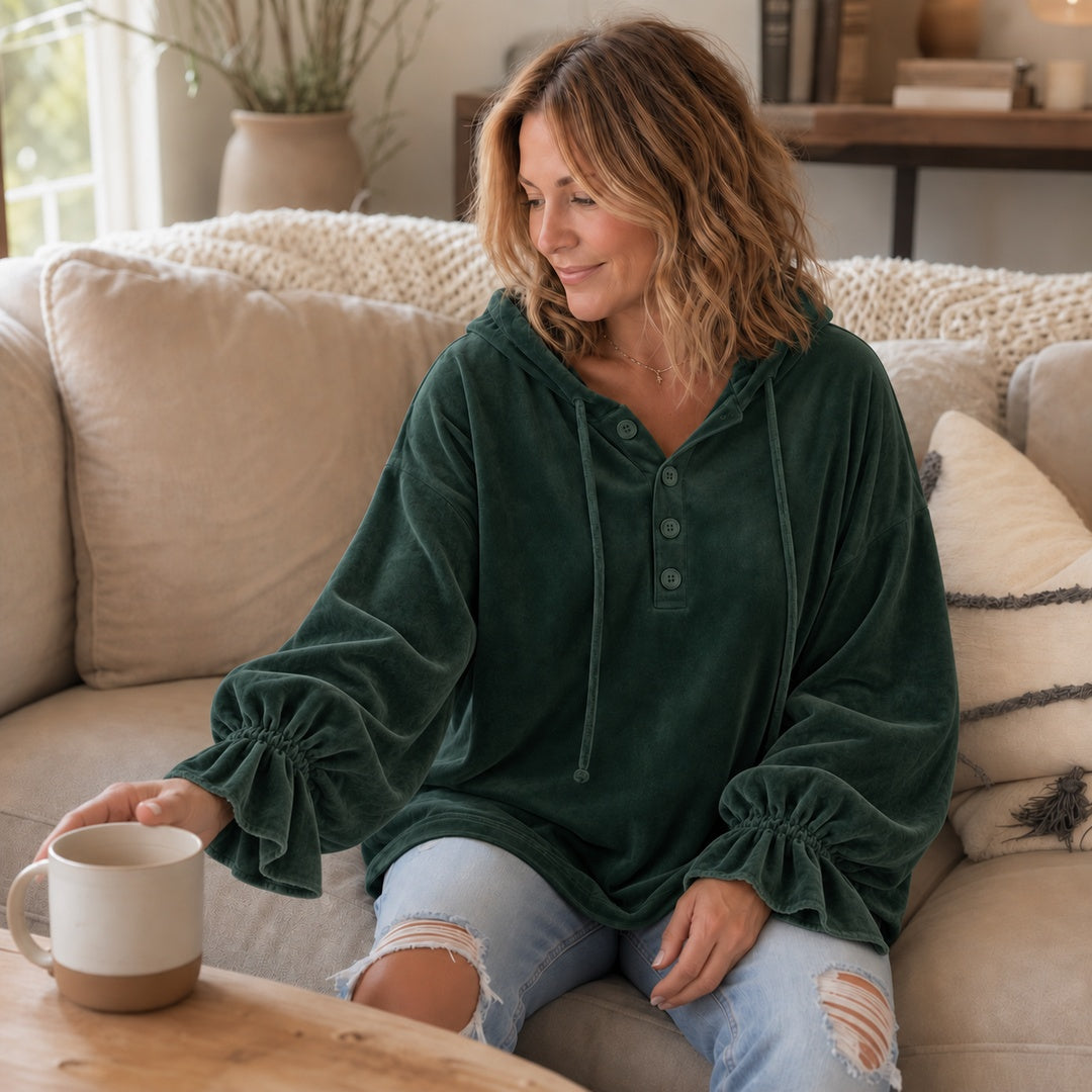 Vrouw in een donkergroene hoodie op de bank, die naar een mok reikt | Boutique Moda
