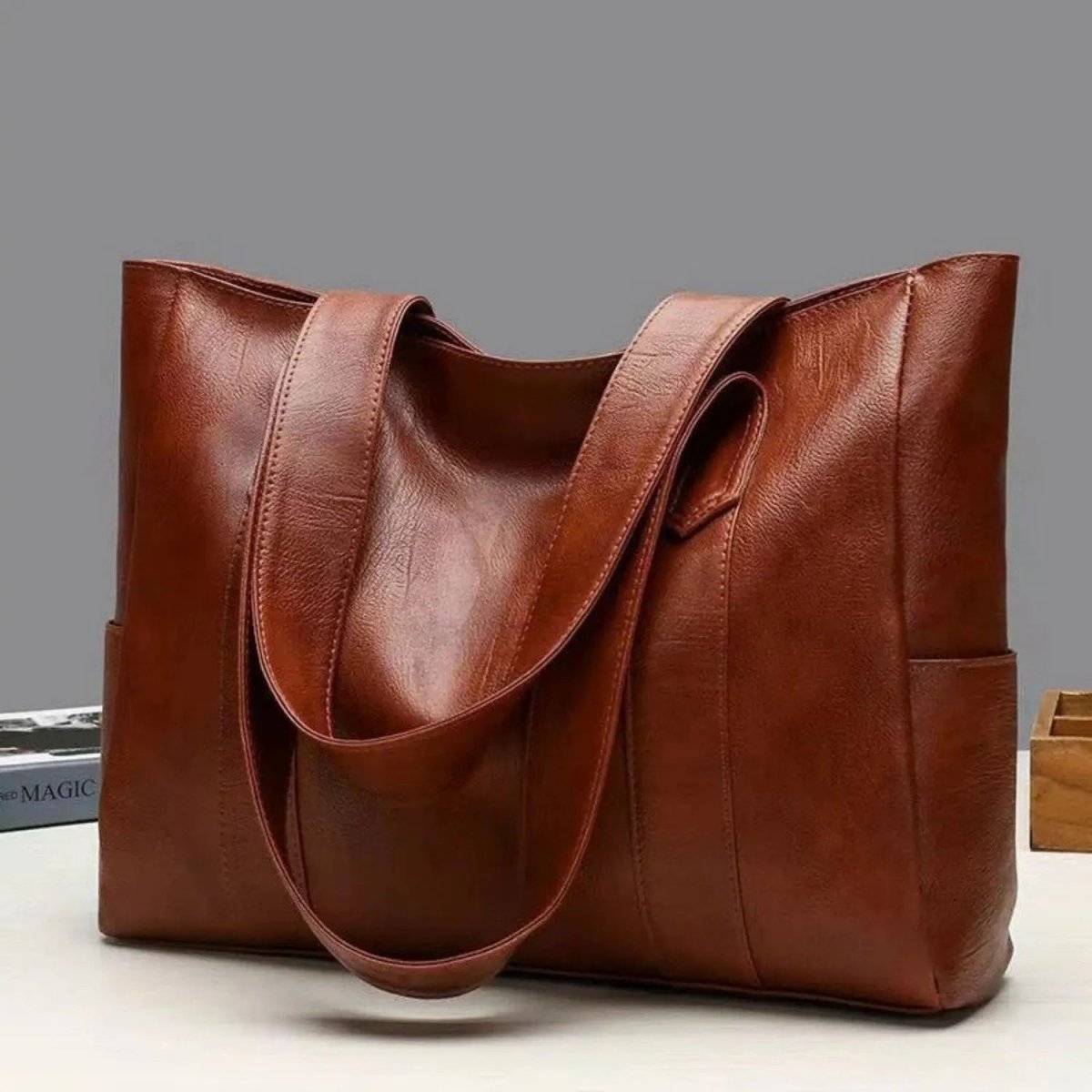 Bruine leren schoudertas met dubbele hengsels, vrijstaand tegen grijze achtergrond | Boutique Moda