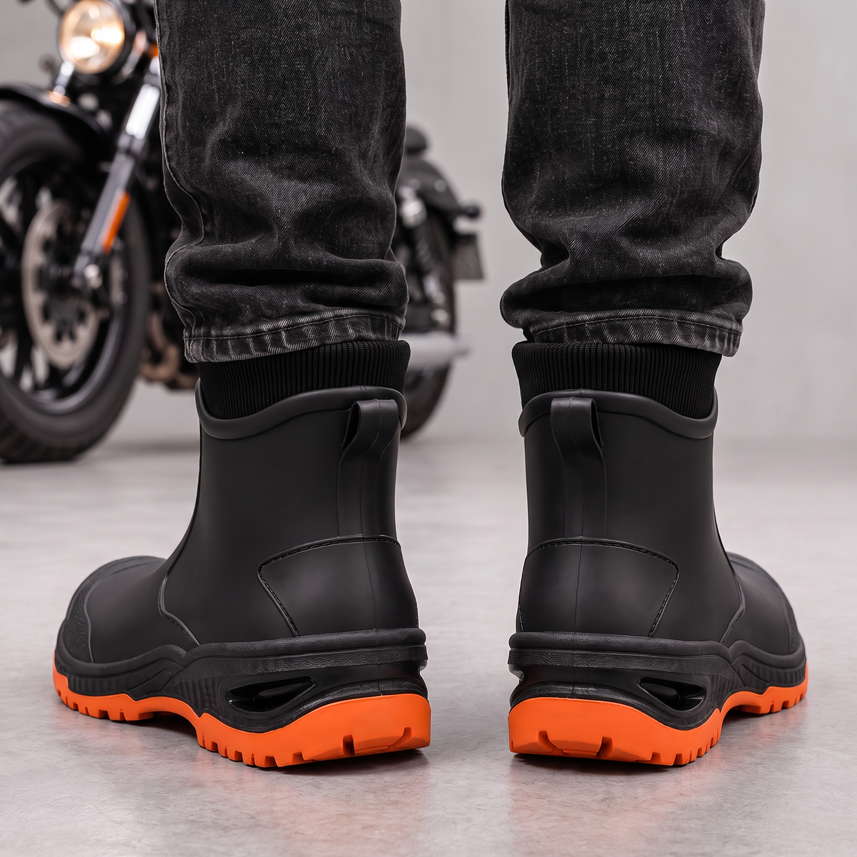 Black ankle rain boots with orange lugged soles worn with dark jeans, back view with pull tabs.